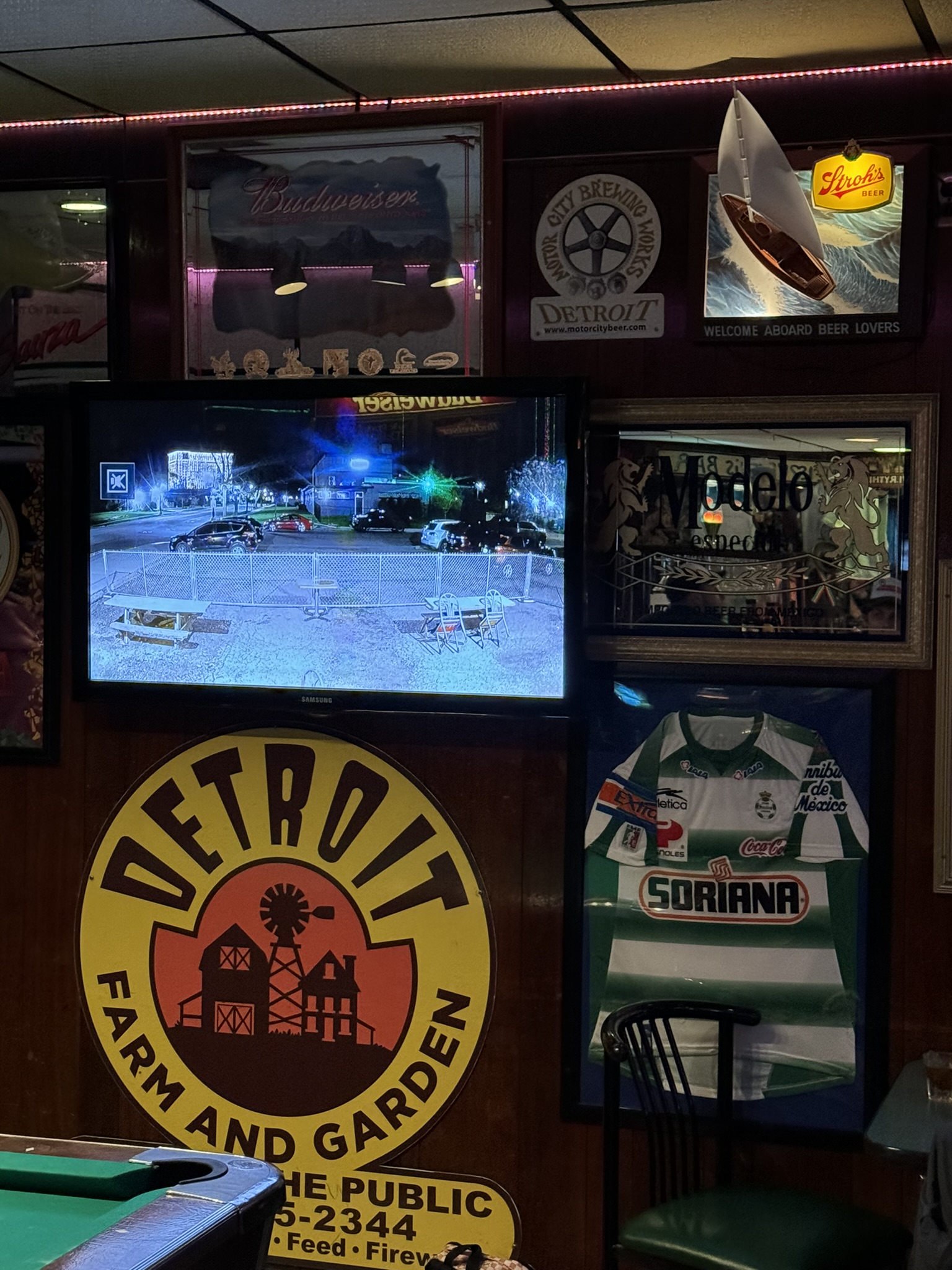 Security monitor live feed of parking lot inside a Detroit dive bar with Budweiser, Stroh’s, and Modelo beer signs next to it. Sign for Detroit Farm and Garden below and Soriana soccer jersey on wall below monitor.