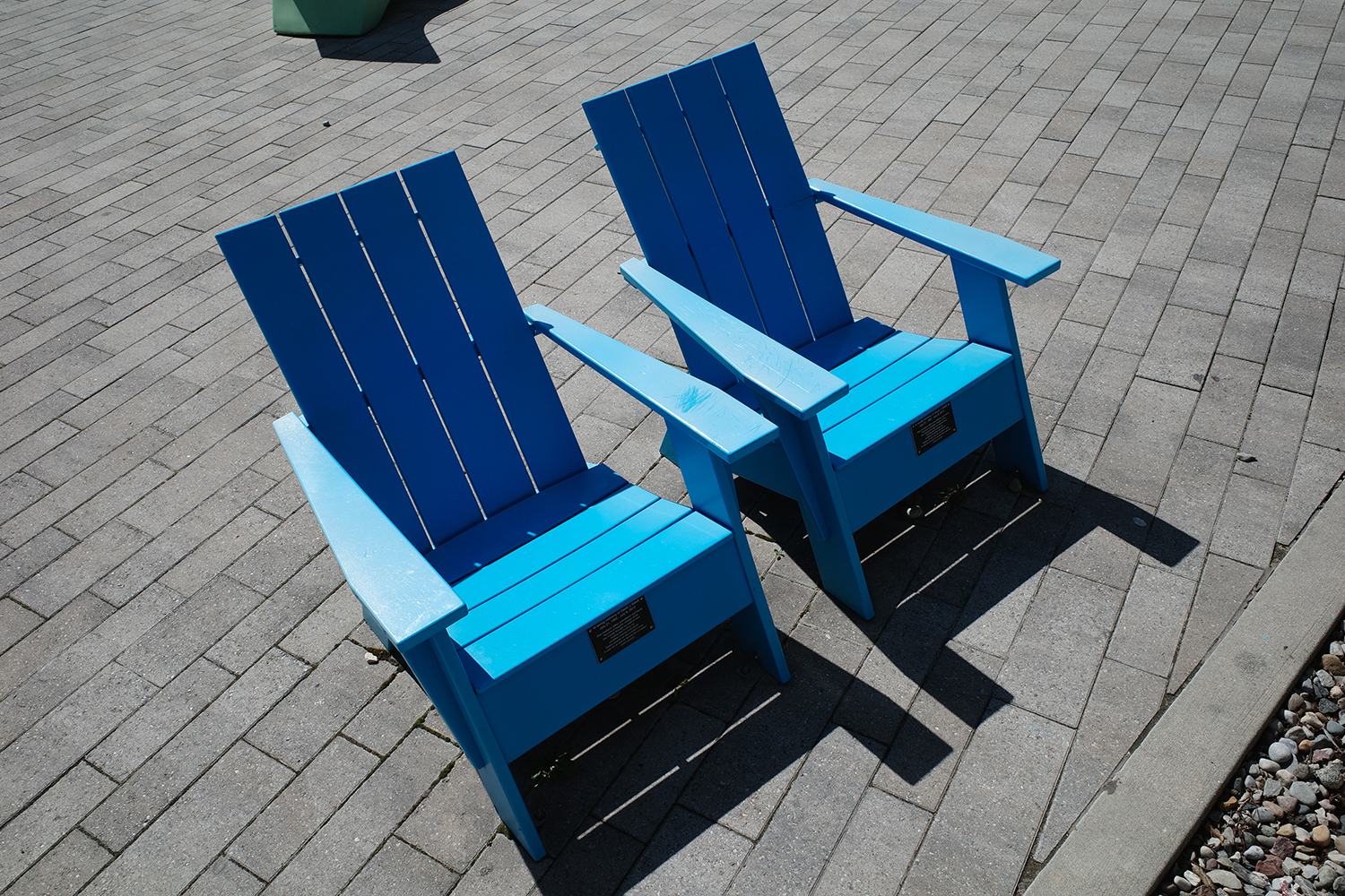 Two blue Adirondack chairs in the sun on a gray stone patio.