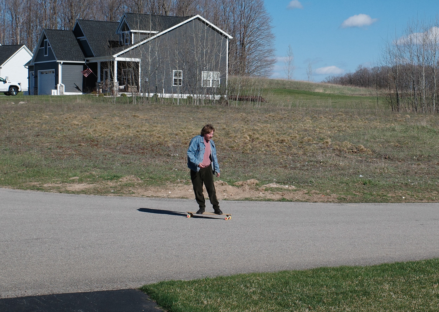 Man riding a longboard on a street in a suburban neighborhood, house with an American flag in the background.