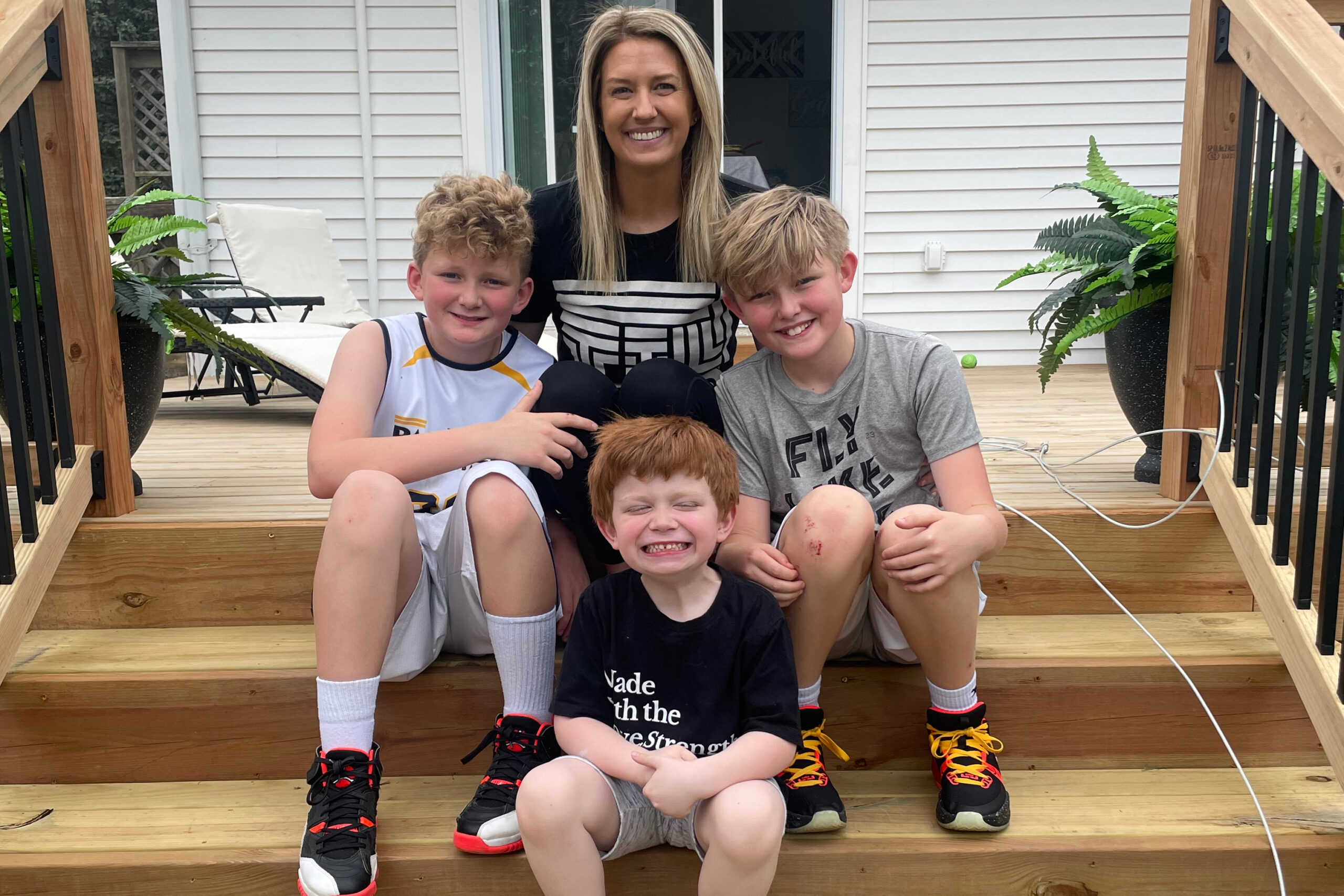 A woman and three small boys sitting on the stairs on a wooden deck outside a home.
