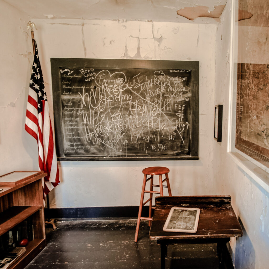 Photo of dilapidated classroom in old schoolhouse in northern Michigan, scribbled illegible text on chalkboard, american flag and bookshelf.