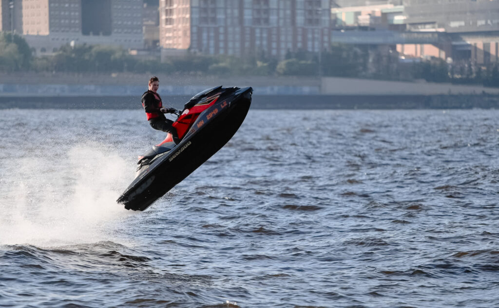 Man on a red jet ski mid-air jumping across a river with a city in the distance.