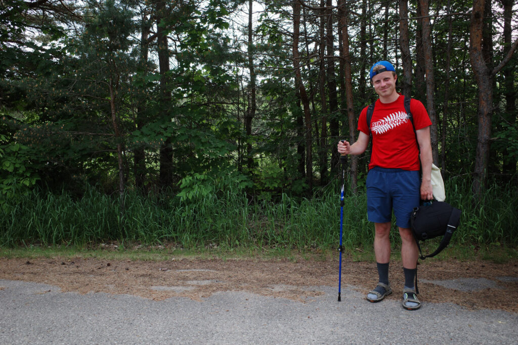 Smiling man in red shirt with hiking poles and sandals standing on pavement road next to forest.