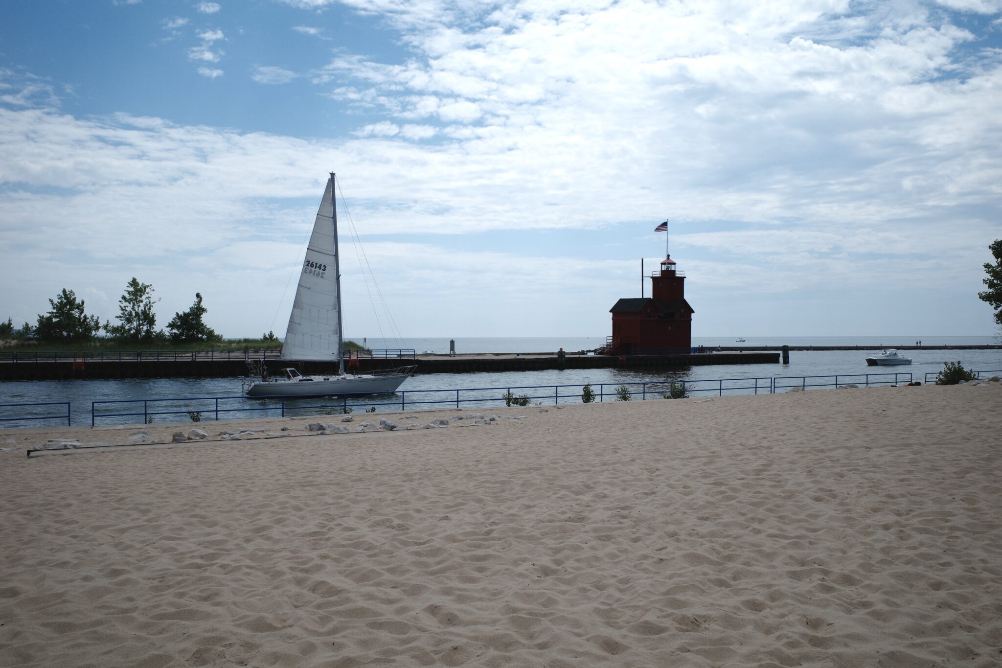 Sailboat traversing an outlet from the beach to lake michigan with red lighthouse in background.