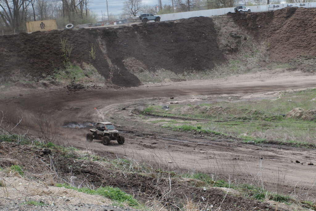 Side by side 4-wheeler speeding around a muddy bend, Jeeps climbing a hill trail in distance.
