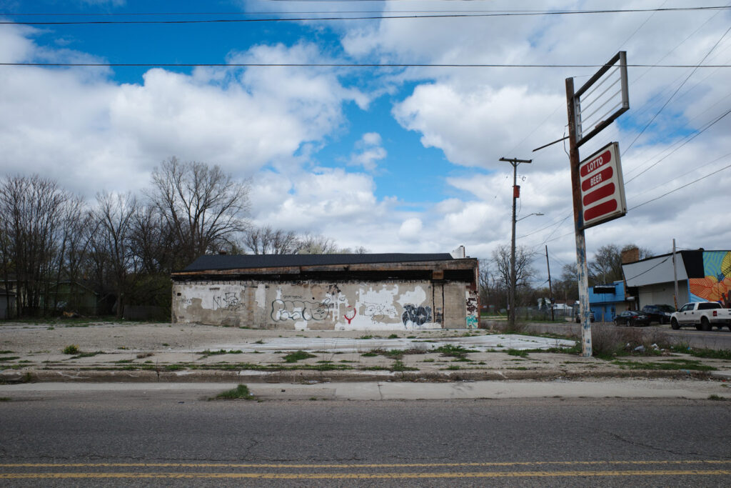 Abandoned convenience store covered in graffiti, parking lot with hollow sign, two remaining parts reading “Lotto” and “Beer.”
