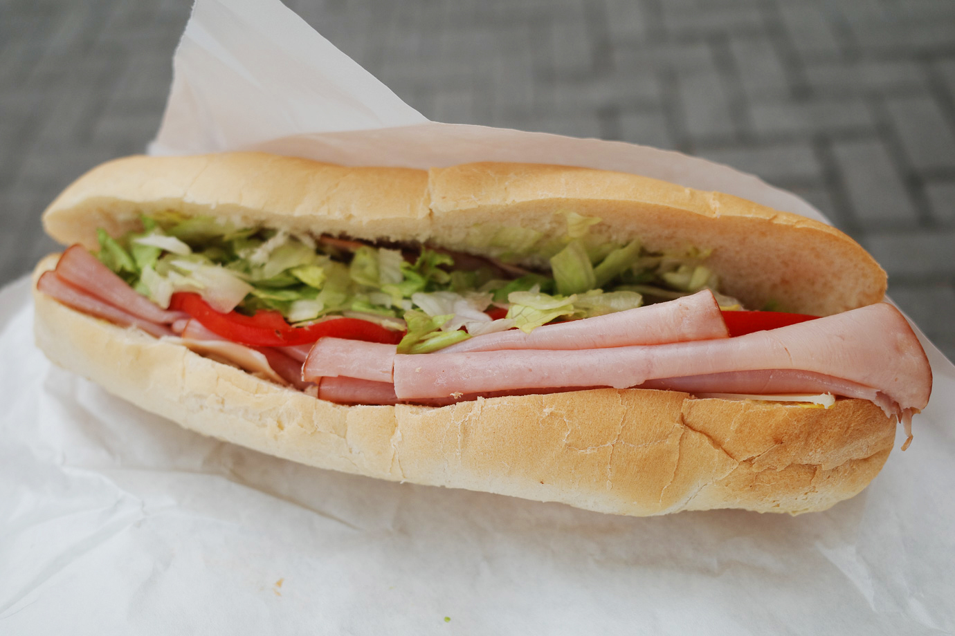 Sub sandwich with ham, cheese, and lettuce on white bread sitting on white paper wrapper.