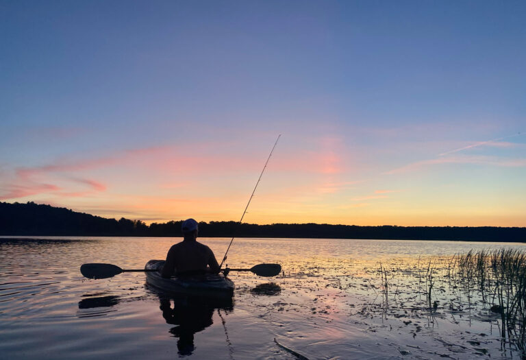 Man in canoe on lake at sunset with fishing reel.