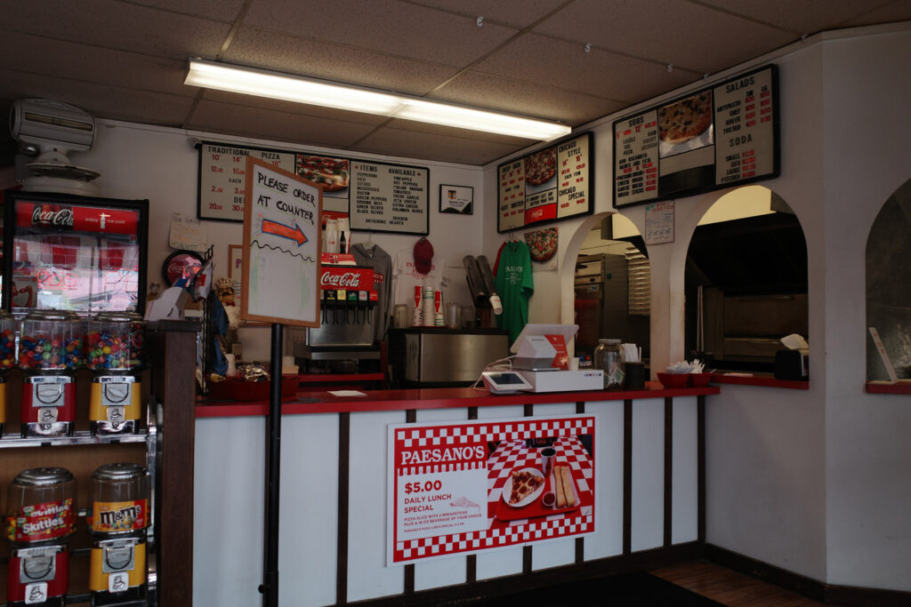 Old style menu signage and red decor, old candy machines and countertop cash register inside Paesano’s restaurant. Signs reading “Paesano’s $5 daily lunch special, pizza slice and 16oz beverage of your choice + two breadsticks” and “Please order at counter”.