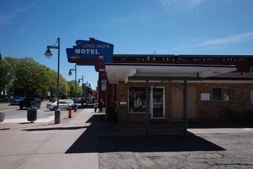 Outside view of Long Ships Motel and its cracked parking lot with vintage sign saying “Long Ships Motel.”