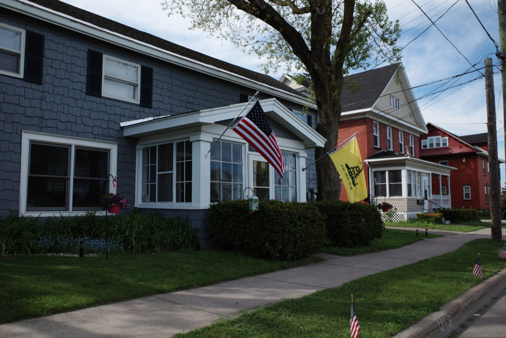 Older residential house on a street with American flag and yellow Gadsden flag displayed out front, small American flags lining the sidewalk.