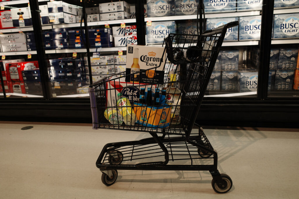 Cart full of Corona Extra, Pabst Blue Ribbon, and liquor bottles near glass cases in grocery store.