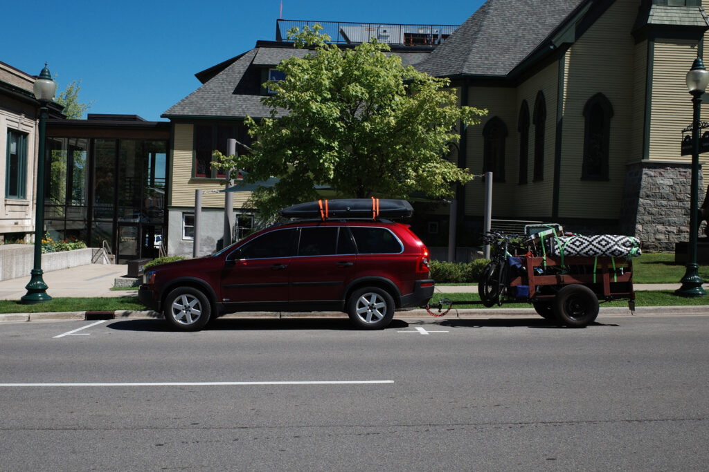 Red SUV with storage unit strapped to roof and trailer holding kayaks and bicycles.