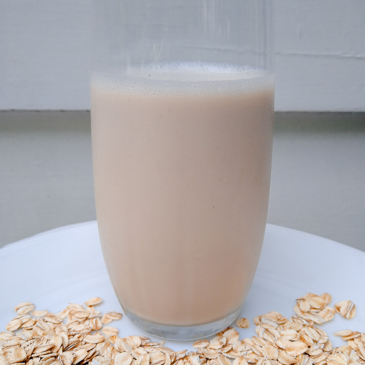 A glass of oat milk on white table with oats in foreground.