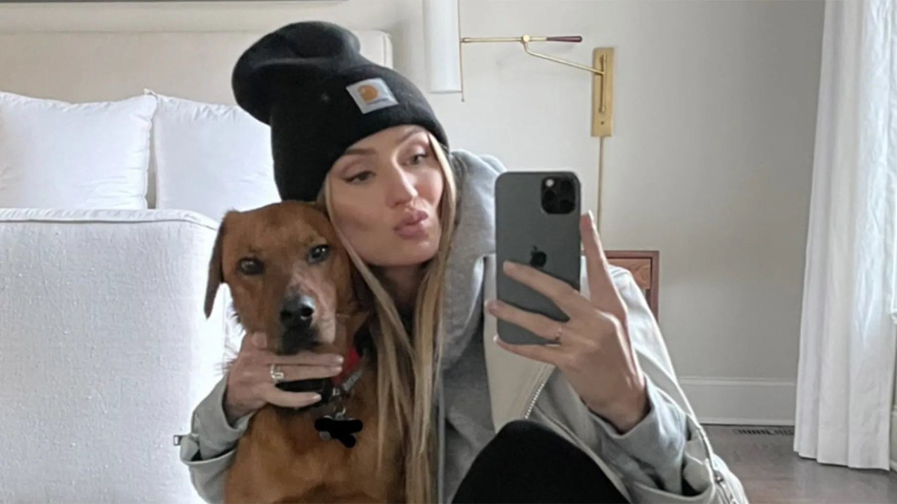 Woman in black carhartt beanie with dog taking mirror selfie.