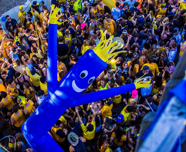 Blue inflatable man smiling with large party crowd in neon gear below.