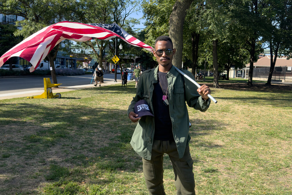 African-American man with aviator sunglasses and Enjoyer hat under his arm and large American flag draped over his shoulder.