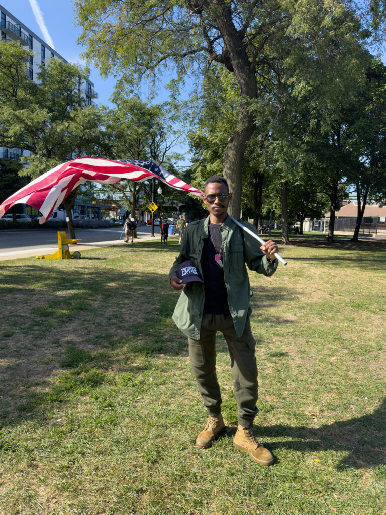 African-American man in aviator sunglasses posing with Enjoyer hat under his arm and large American flag draped over shoulder.