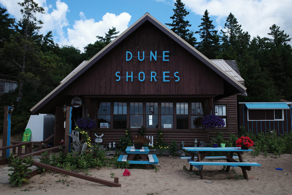 Dune Shores pasty restaurant, a cabin with dark wood and benches outside, with sign saying "Dune Shores"