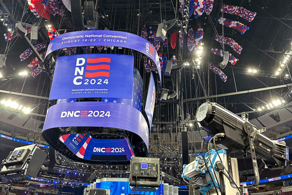 Inside of the DNC with signs reading "Democratic National Convention August 19-22 Chicago" and "DNC 2024." with balloons held in nets at ceiling and TV cameras in foreground.