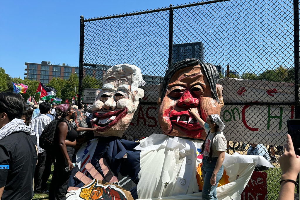 Caricature masks of Joe Biden and Kamala Harris with fangs dripping blood being painted, next to asian caricature on the ground.