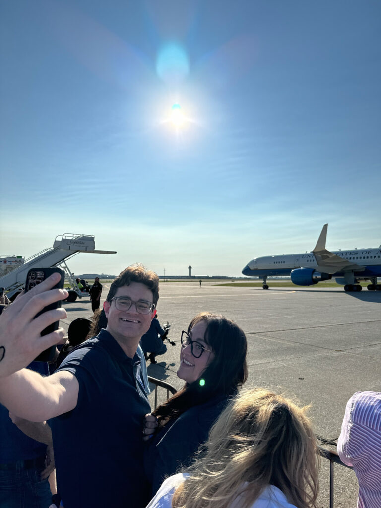 Man holding up phone to take selfie with woman with Air Force 2 on tarmac in background.