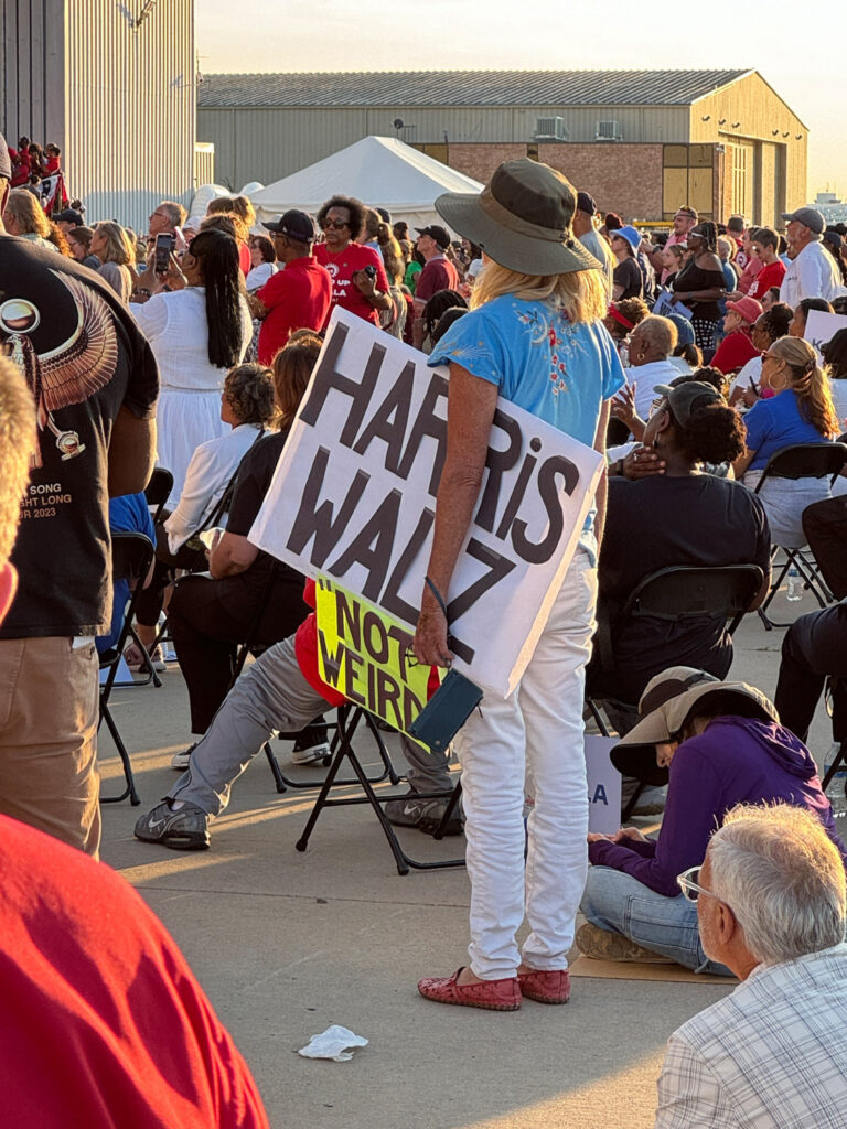 Woman holding sign "Harris Walz Not Weird" outside hangar in small crowd at Kamala Harris rally.