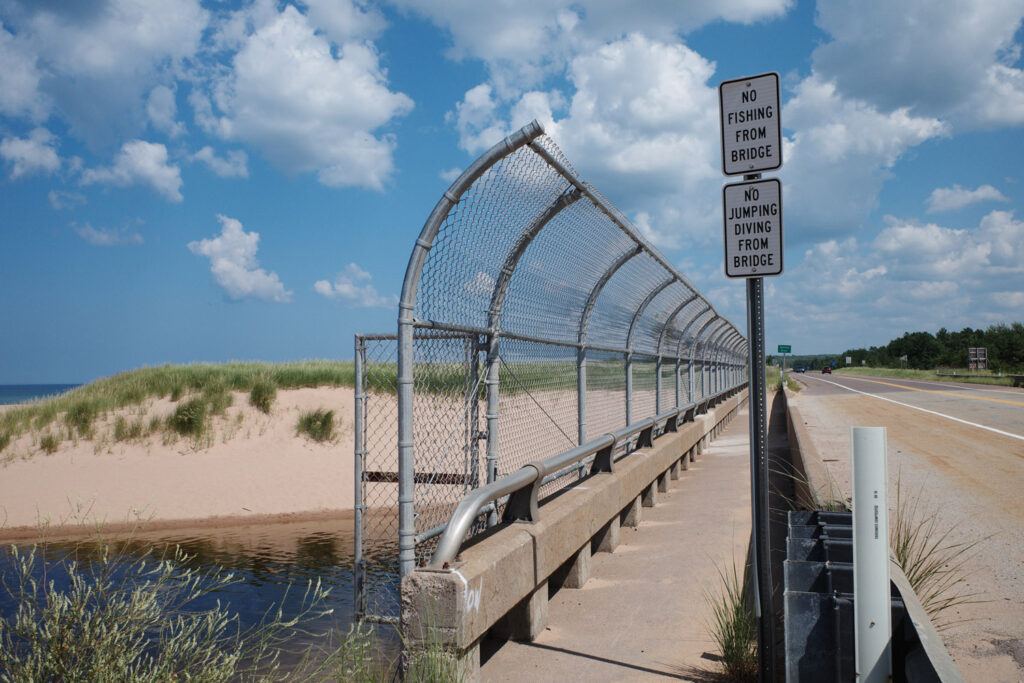 Chainlink fence on bridge over river with signs reading, "no fishing from bridge" and "no jumping diving from bridge."