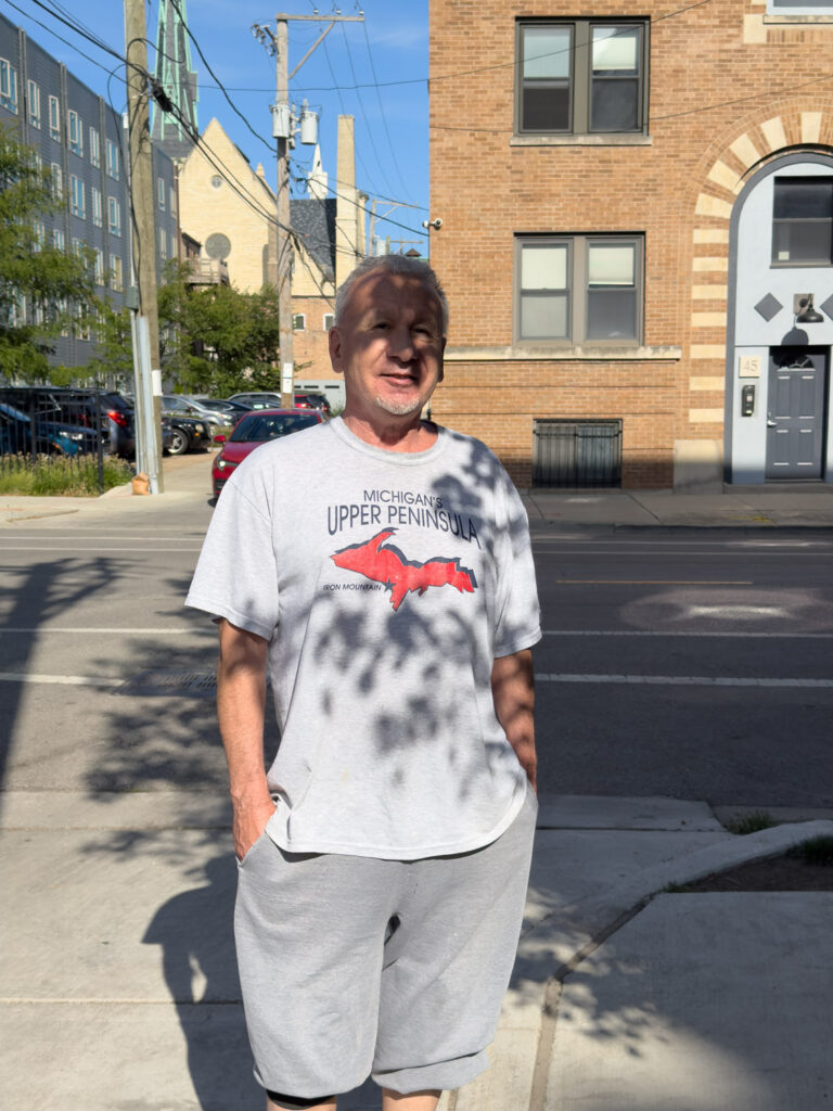 Older white man in shirt reading "Michigan's Upper Peninsula, Iron Mountain" on street.