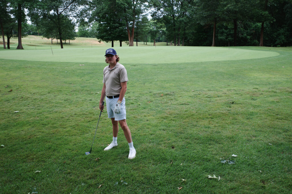 Bobby Mars in Enjoyer hat with chipping wedge next to practice green.