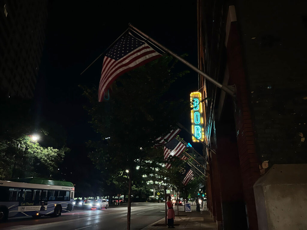 Outside of the B.O.B. comedy club with B.O.B. light up sign and american flags hanging, person on street and car traffic.