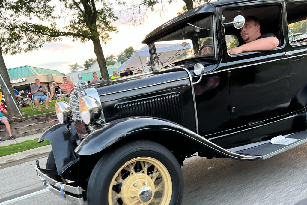 1920s style car racing down street with onlookers on side of road.