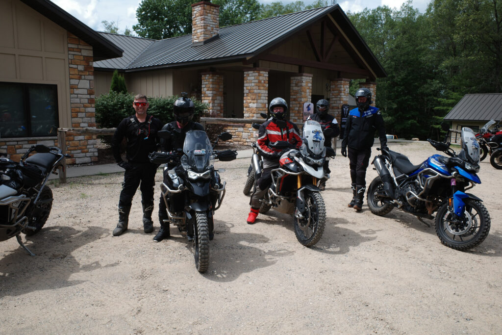 Men smiling next to motorcycles in dirt parking lot outside building flanked by trees.