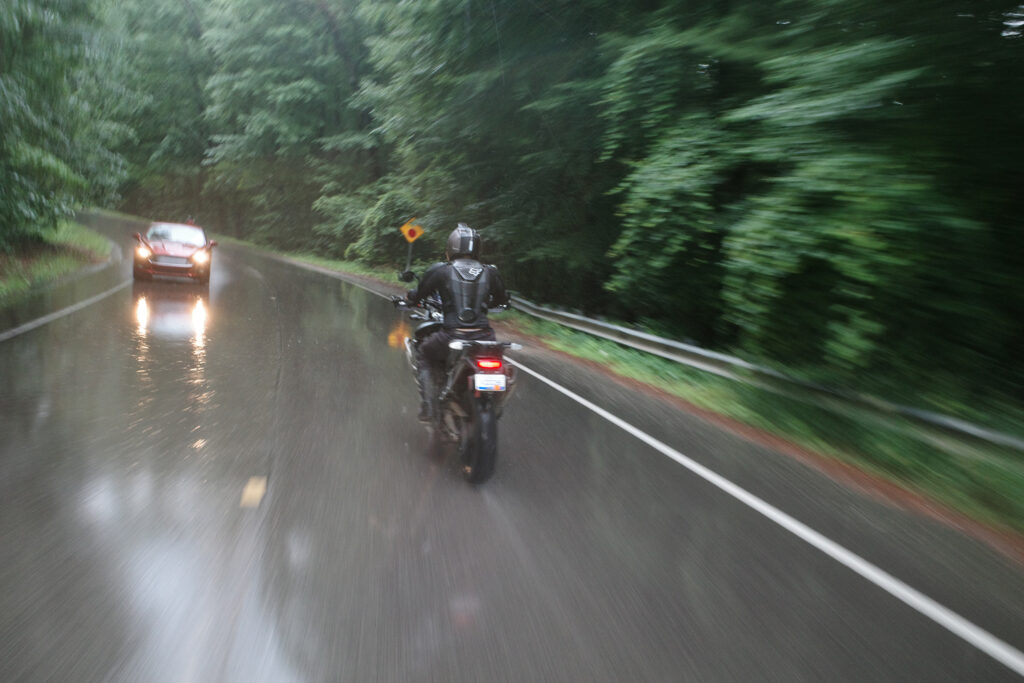 Man riding motorcycle on wet pavement street in forest as car approaches in opposite lane.