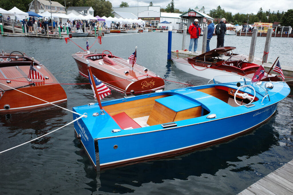 Wooden boats docked, one painted blue, one named "Ruby," men gathering on dock with tents and crowd in background.