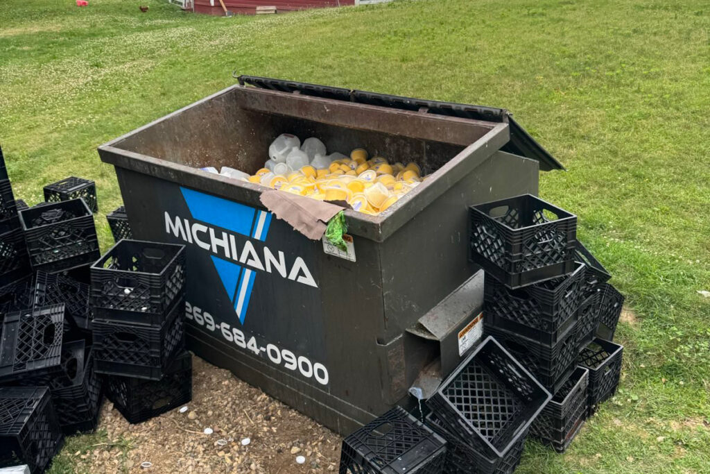 Pile of raw milk products sitting in dumpster labeled "Michiana" with phone number 269-694-0900 with farm in background.