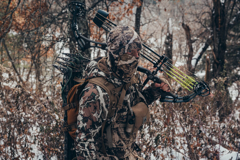 Man in winter scene dressed in winter camo and balaclava with bow slung over his shoulder.