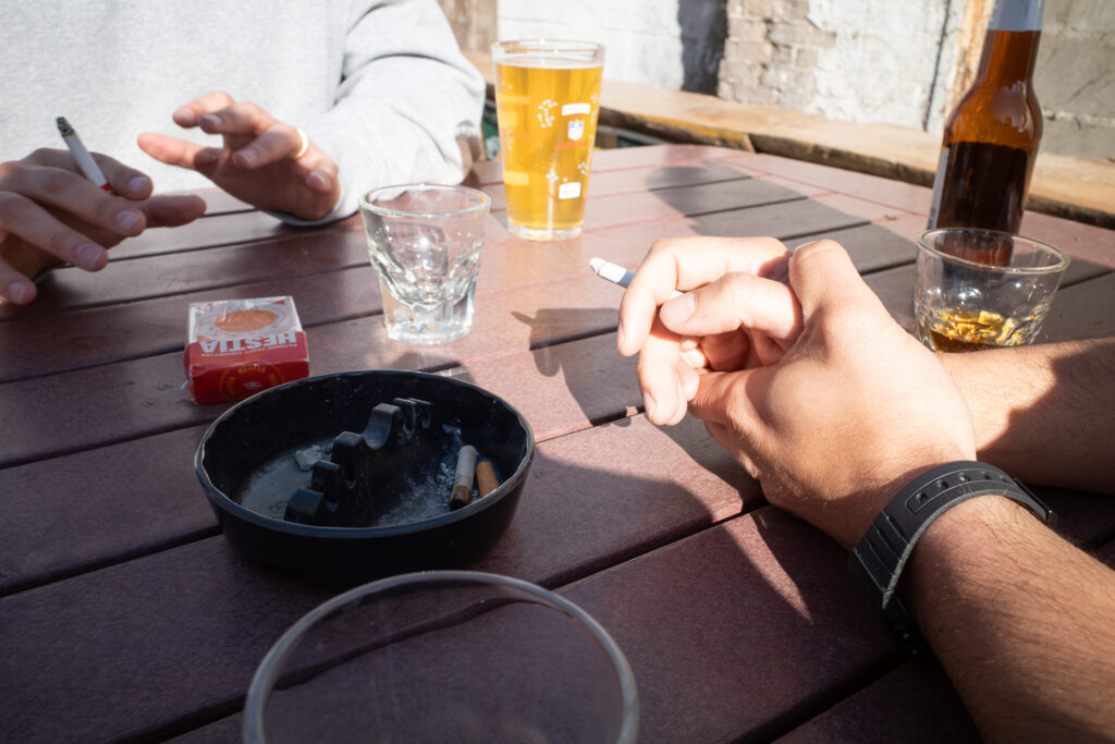 Men's hands on outdoor table in sun with drinks and cigarettes.