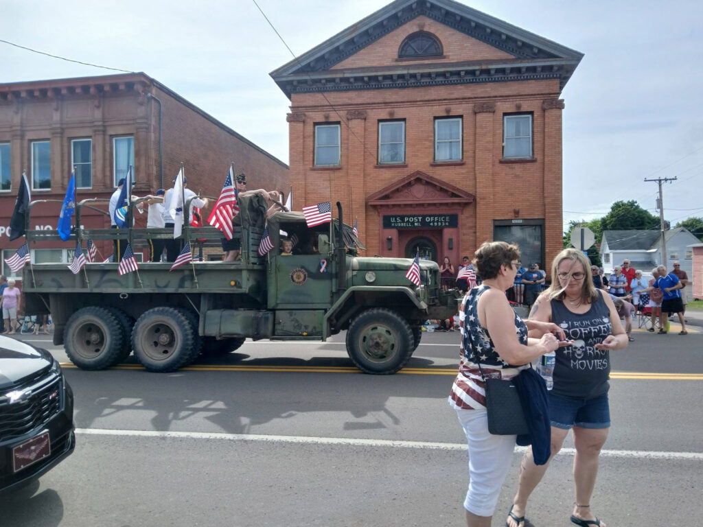 Military truck during parade on street with American flags out front of old U.S. Post Office.