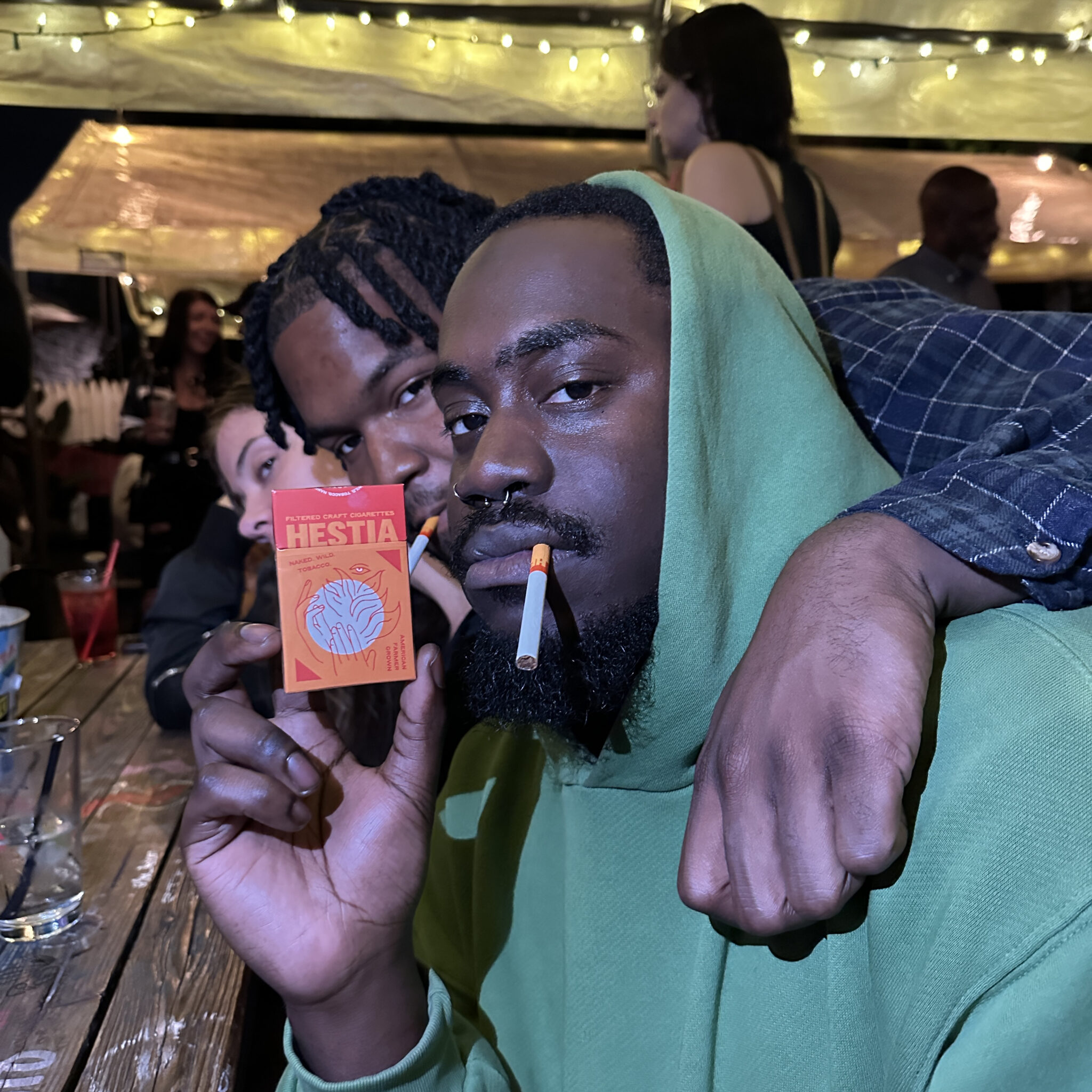 Two men smoking and posing with Hestia cigarettes box.