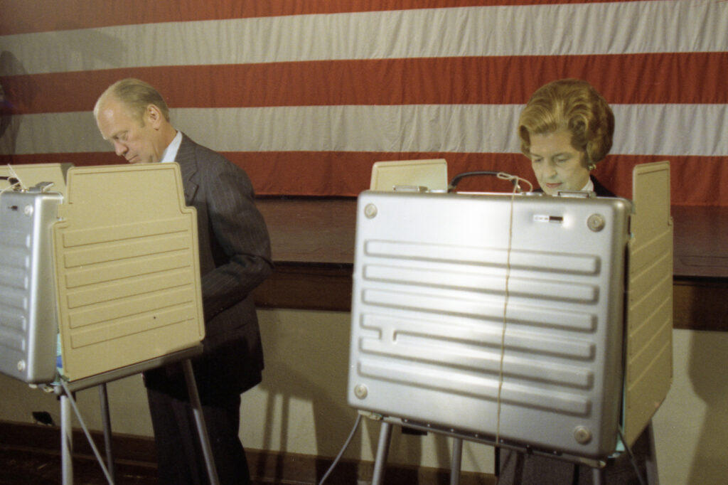 President Gerald Ford and First Lady Betty Ford in voting booths with American flag in background.