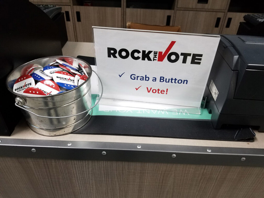 Rock the vote sign also reading "grab a button" and "vote" with "I voted" buttons in bucket.