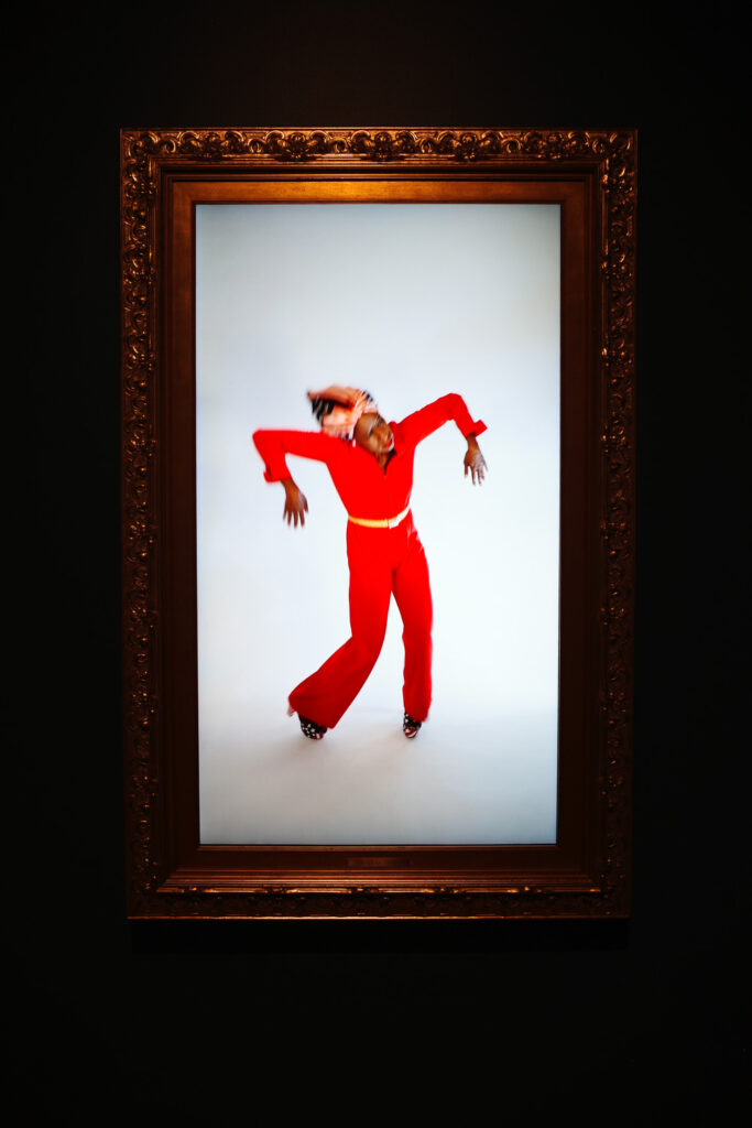 Black woman in red outfit dancing in video, screen housed in an ornate frame.