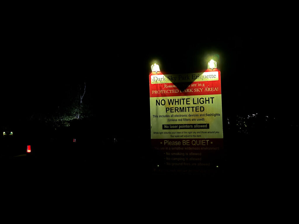 Lit up sign at night reading "Dark Sky Park Etiquette Remember you are in a protected dark sky area! no white light permitted this includes all electronic devices and flashlights (unless red filters are used) no laser pointers allowed. White light disturbs your view of the night sky and those around you. Your eyes will adjust to the dark. Please be quiet. No smoking is allowed."