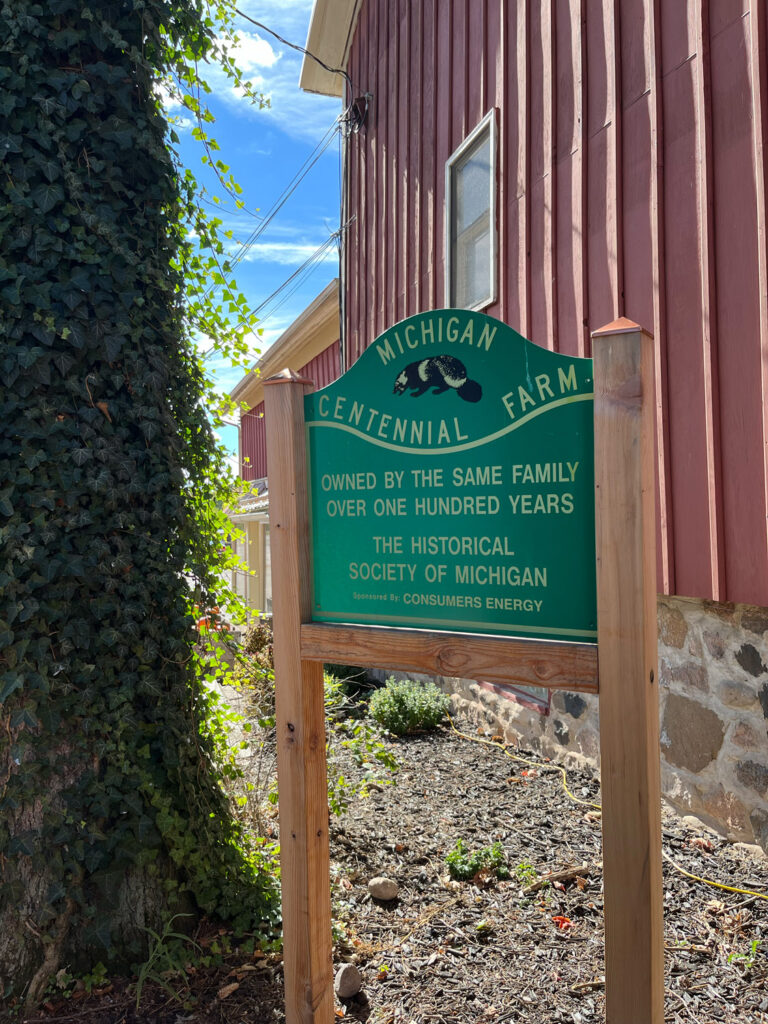 Sign outdoors reading "Michigan Centennial Farm, Owned by the Same Family Over One Hundred Years, The Historical Society of Michigan Sponsored by Consumers Energy"