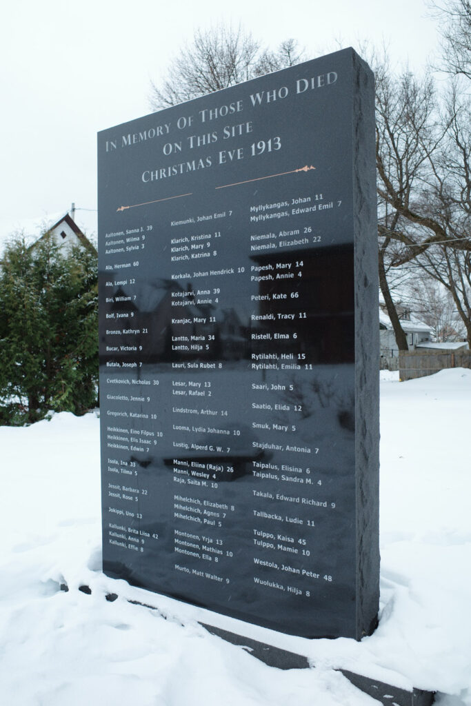 Commemorative plaque reading "IN MEMORY OF THOSE WHO DIED
ON THIS SITE
CHRISTMAS EVE 1913
Aaltonen, Sanna J. 39
Aaltonen, Wilma 9
Aaltonen, Sylvia 3
Ala, Herman 60
Ala, Lempi 12
Biri, William 7
Bolf, Ivana 9
Bronzo, Kathryn 21
Bucar, Victoria 9
Butala, Joseph 7
Cvetkovich, Nicholas 30
Giacoletto, Jennie 9
Gregorich, Katarina 10
Heikkinen, Eino Filpus 10
Heikkinen, Elis Isaac 9
Heikkinen, Edwin 7
Isola, Ina 33
Isola, Tilma 5
Jessit, Barbara 22
Jessit, Rose S
Jokippi, Uno 13
Kallunki, Brita Liisa 42
Kallunki, Anna 9
Kallunki, Effie 8
Kiemunki, Johan Emil 7
Klarich, Kristina 11
Klarich, Mary 9
Klarich, Katrina 8
Myllykangas, Johan 11
Myllykangas, Edward Emil 7
Niemala, Abram 26
Niemala, Elizabeth 22
Korkala, Johan Hendrick 10
Kotajarvi, Anna 39
Kotajarvi, Annie 4
Papesh, Mary 14
Papesh, Annie 4
Peteri, Kate 66
Renaldi, Tracy 11
Kranjac, Mary 11
Ristell, Elma 6
Lantto, Maria 34
Lantto, Hilja 5
Lauri, Sula Rubet 8
Rytilahti, Heli 15
/Rytilahti, Emilia 11
Lesar, Mary 13
Lesar, Rafael 2
Saari, John 5
Saatio, Elida 12
Lindstrom, Arthur 14
Smuk, Mary 5
Luoma, Lydia Johanna 10
Lustig, Alperd G. W. 7
Stajduhar, Antonia 7
Manni, Elina (Raja) 26
Manni, Wesley 4
Raja, Saita M. 10
Taipalus, Elisina 6
Taipalus, Sandra M. 4
Takala, Edward Richard 9
Mihelchich, Elizabeth 8
Mihelchich, Agnes 7
Mihelchich, Paul 5
Montonen, Yrja 13
Montonen, Mathias 10
Montonen, Ella 8
Murto, Matt Walter 9
Tallbacka, Ludie 11
Tulppo, Kaisa 45
Tulppo, Mamie 10
Westola, Johan Peter 48
Wuolukka, Hilja 8"