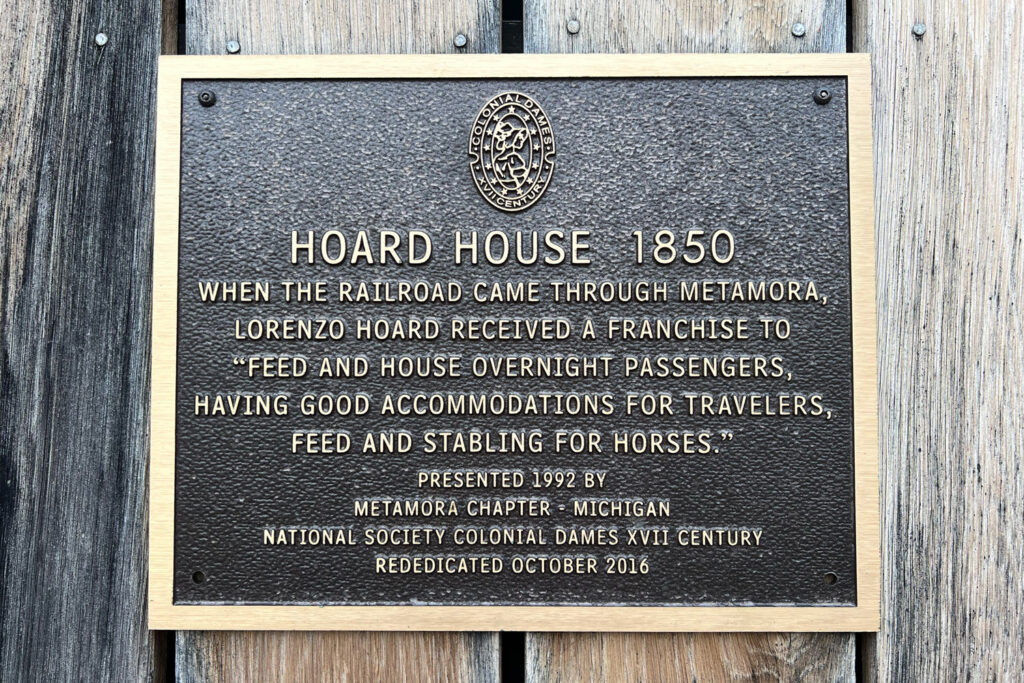 plaque reading "HOARD HOUSE 1850
WHEN THE RAILROAD CAME THROUGH METAMORA, LORENZO HOARD RECEIVED A FRANCHISE TO FEED AND HOUSE OVERNIGHT PASSENGERS, HAVING GOOD ACCOMMODATIONS FOR TRAVELERS, FEED AND STABLING FOR HORSES." PRESENTED 1992 BÝ
METAMORA CHAPTER = MICHIGAN
NATIONAL SOCIETY COLONIAL DAMES XVII CENTURY
REDEDICATED OCTOBER 2016"