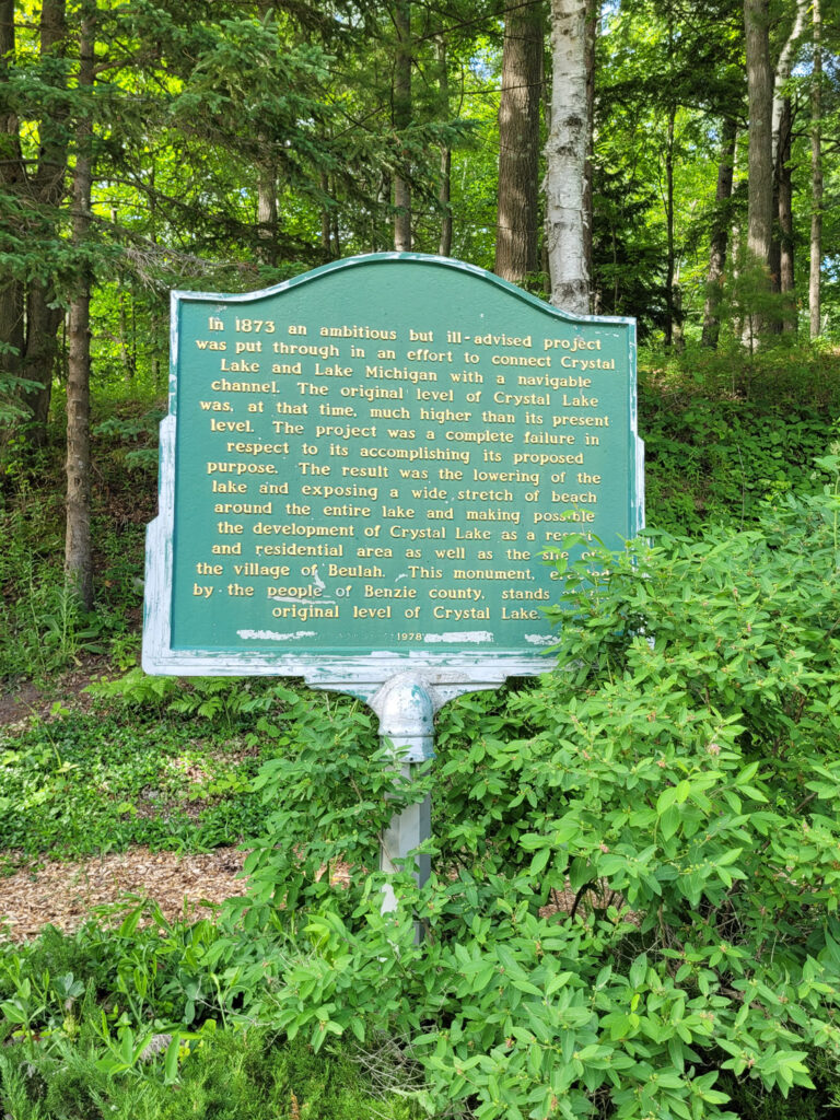 sign reading "In
1873 an ambitious but ill-advised project was put through in an effort to connect Crystal Lake and Lake Michigan with a navigable Channel. The original level of Crystal Lake was, at that time, much higher than its present level.
The project was a complete failure in respect to its accomplishing its proposed purpose. The result was the lowering of the lake and exposing a wide, stretch of beach around the entire lake and making possible the development of Crystal Lake as a res and residential area as well as the sie the village of Beulah. This monument, es by the people of Benzie county, stands
original level of Crystal Lake"