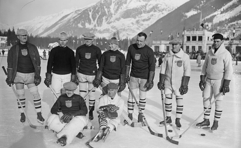 A black and white photo of a group of hockey players wearing matching uniforms with a shield logo, standing and sitting outdoors in a snowy setting, the 1924 U.S. Olympic hockey team.
