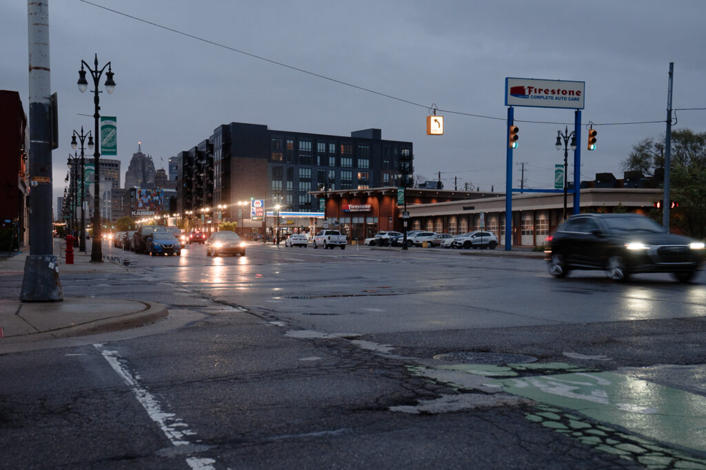 A nighttime urban street scene with a Firestone auto care shop, several vehicles, and buildings with illuminated windows in the background.